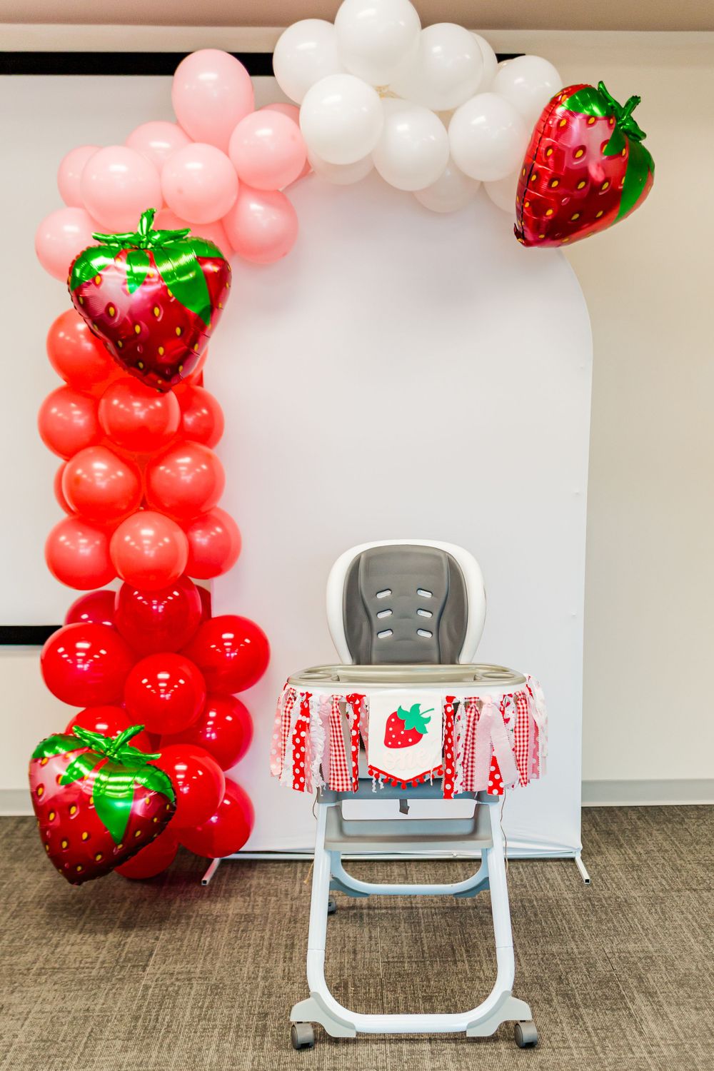 Balloon arch with strawberry decorations and a high chair for a first birthday party.