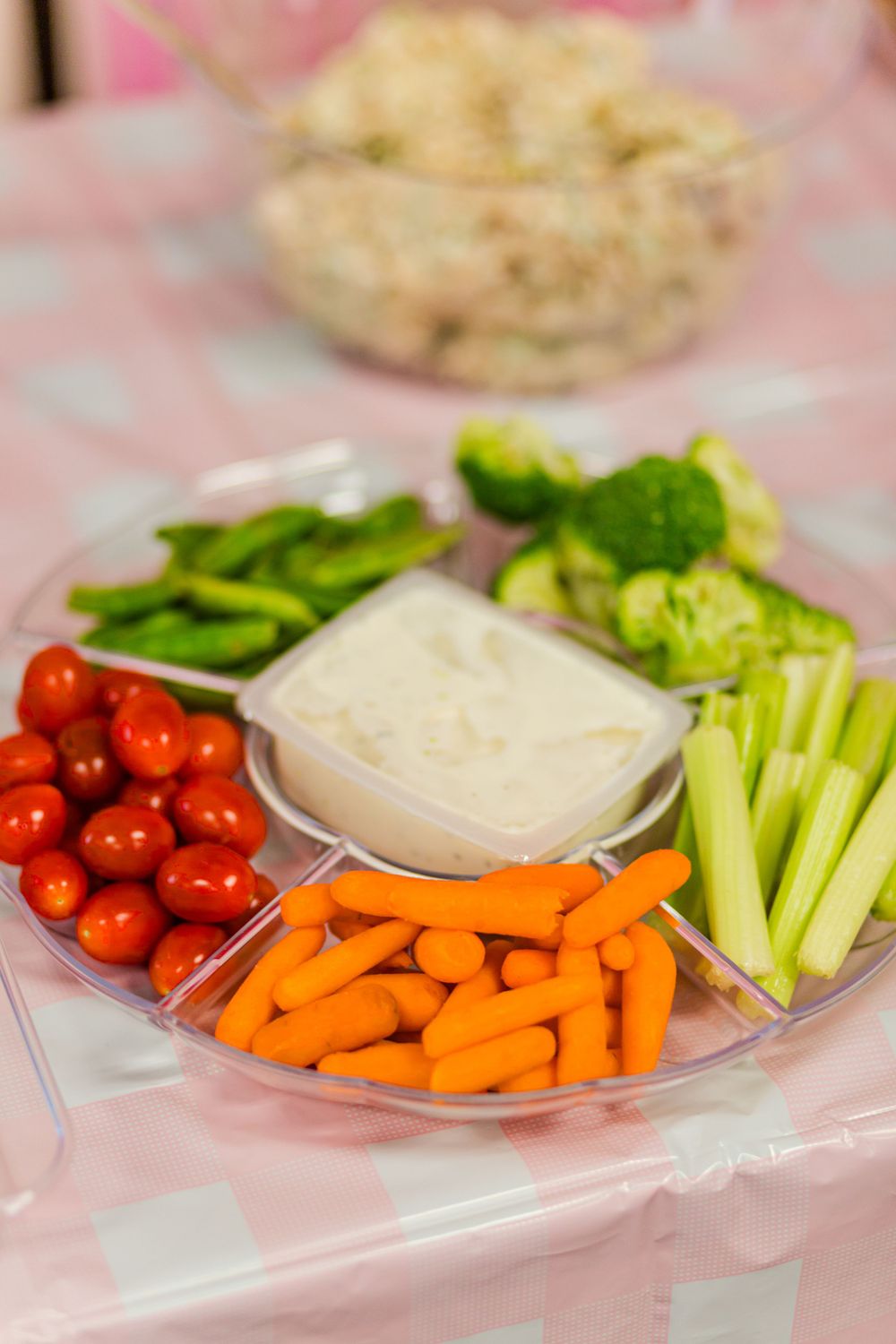 Fresh vegetable platter with carrots, celery, tomatoes and ranch dip on a pink tablecloth.