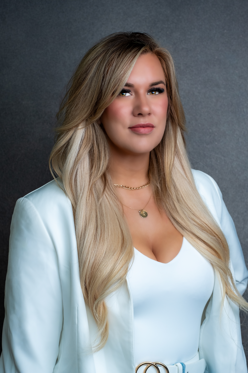 Professional portrait photo of a person wearing a white blazer and top with long blonde hair against a dark grey background.