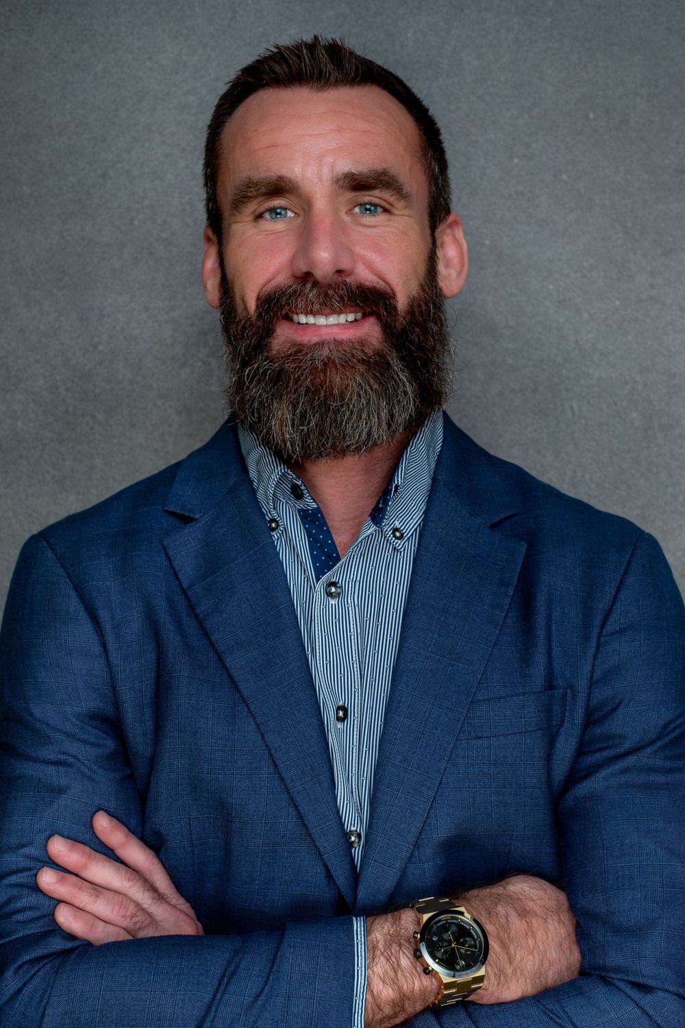 Professional headshot of someone in a navy suit and striped shirt wearing a gold watch and sporting a full dark beard.