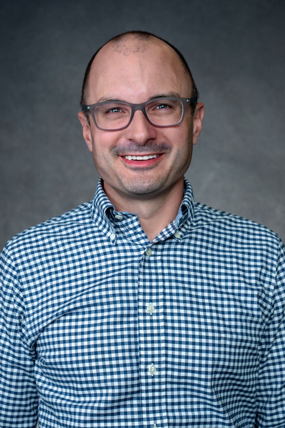 Professional headshot of someone in glasses and a blue gingham button-down shirt against a gray background.