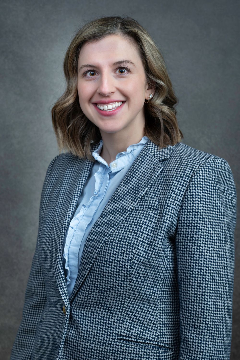 Professional headshot portrait of someone wearing a blue button-down shirt and gray houndstooth blazer against gray background.