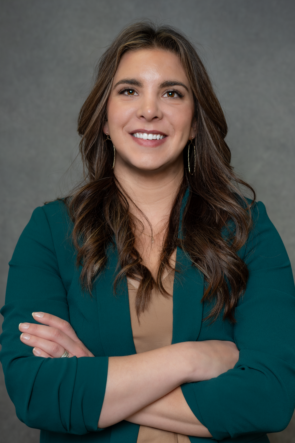 Professional headshot portrait of someone wearing a teal blazer and beige top against a gray backdrop.