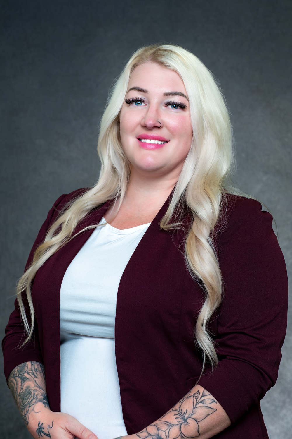 Professional studio portrait of a person with long blonde hair wearing a burgundy blazer and white top with visible arm tattoos.