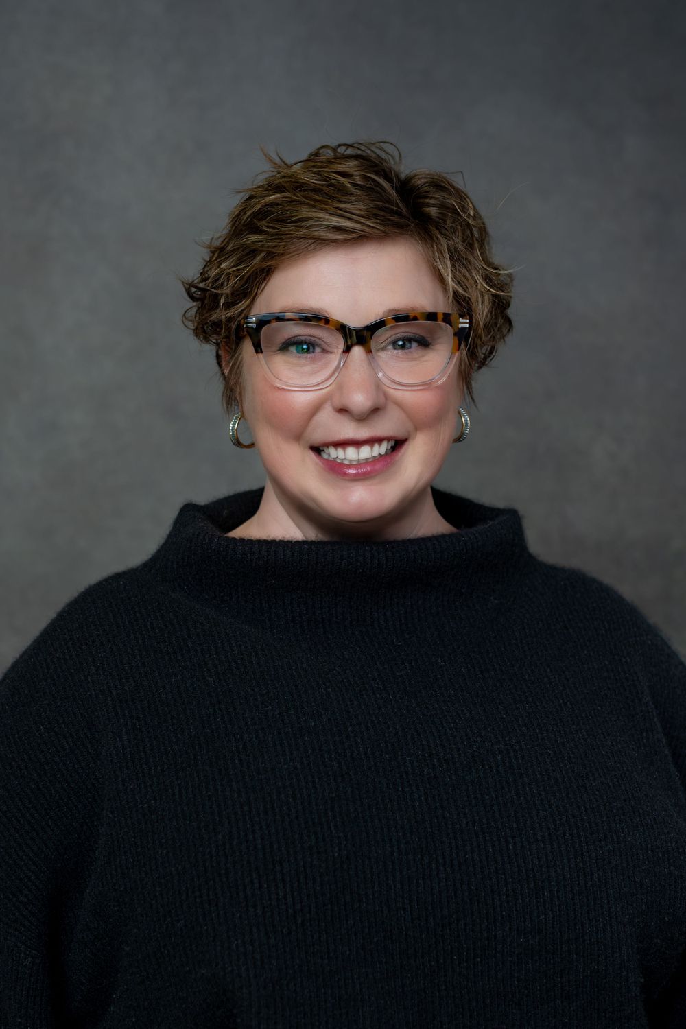 Professional headshot of someone wearing glasses and black sweater smiling warmly against gray background.
