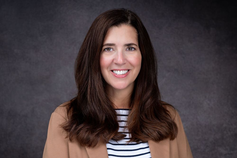 Professional headshot of someone wearing a tan blazer and striped shirt against a dark gray background.