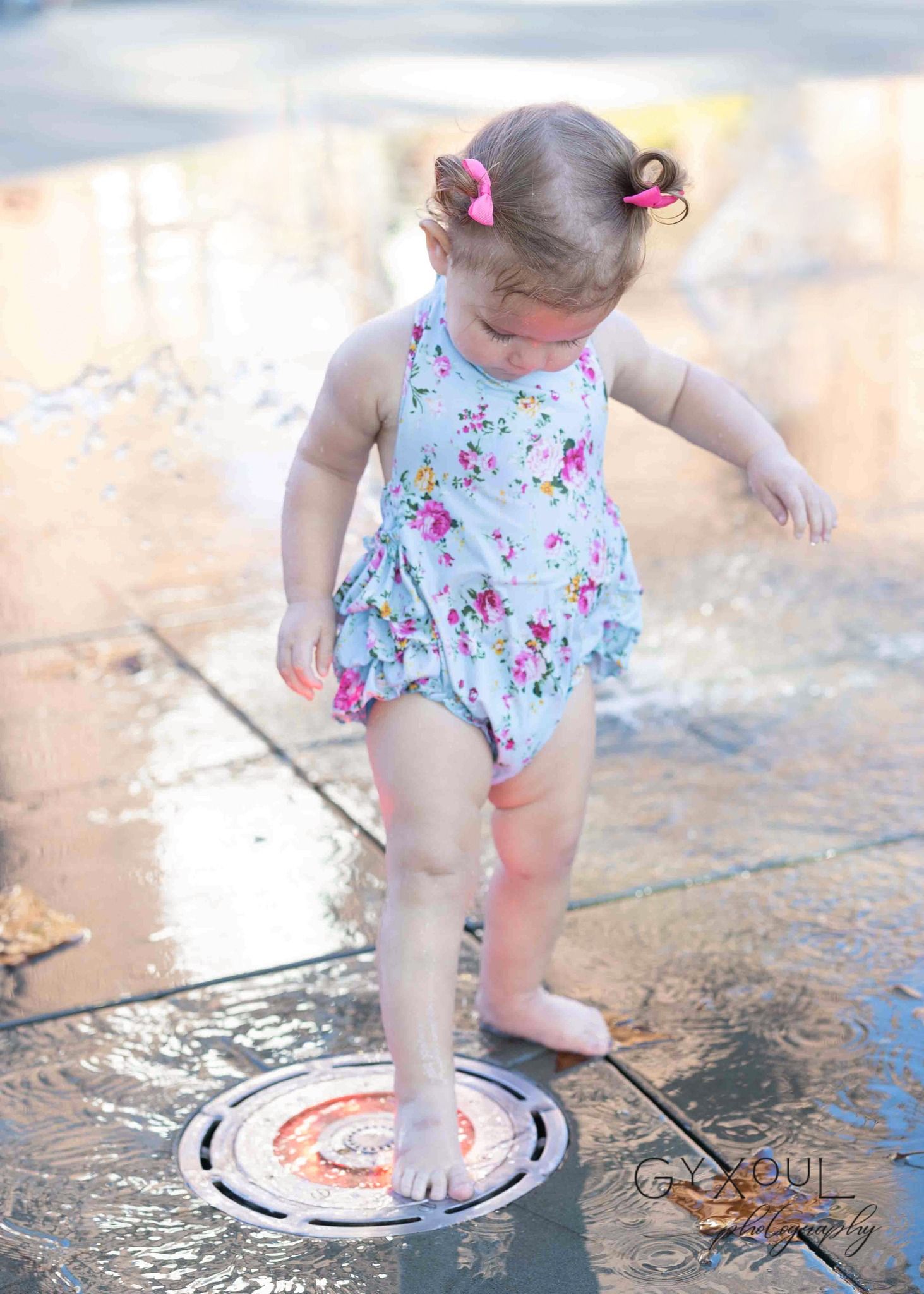 Small child in floral swimsuit playing in water splash pad on sunny day.