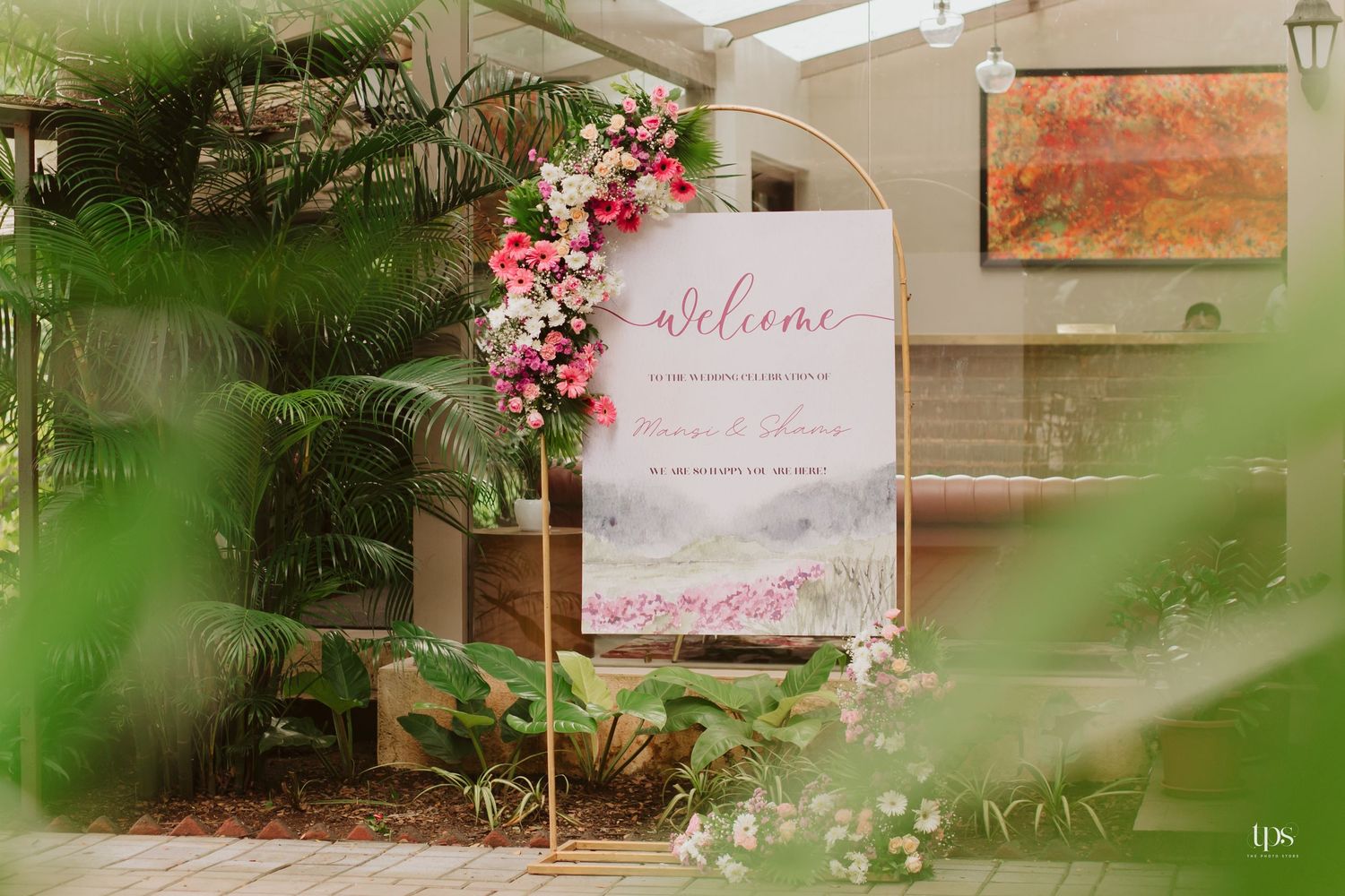 Welcome sign with pink floral decorations framed by tropical plants and greenery.