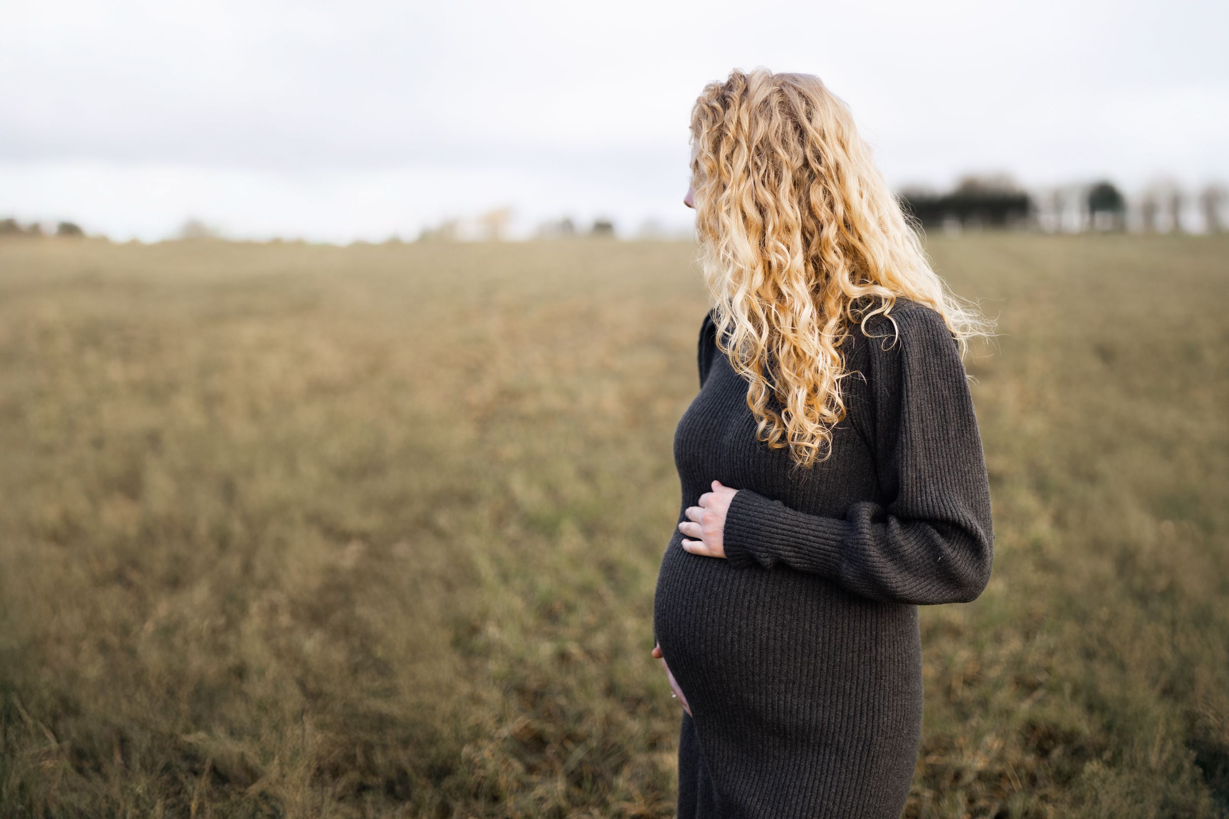 Person with long blonde curly hair wearing a dark sweater stands thoughtfully in an autumn field.