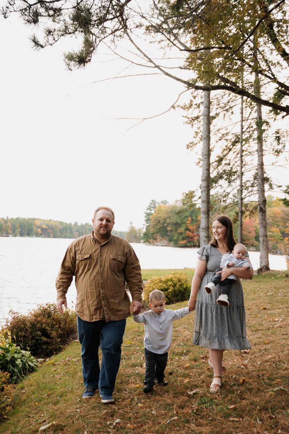 A family walks together on a sloping lakeside trail during autumn with colorful fall foliage in the background.