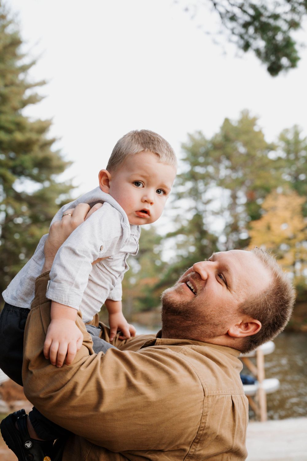 Parent lifting baby in white shirt against backdrop of pine trees near lake.