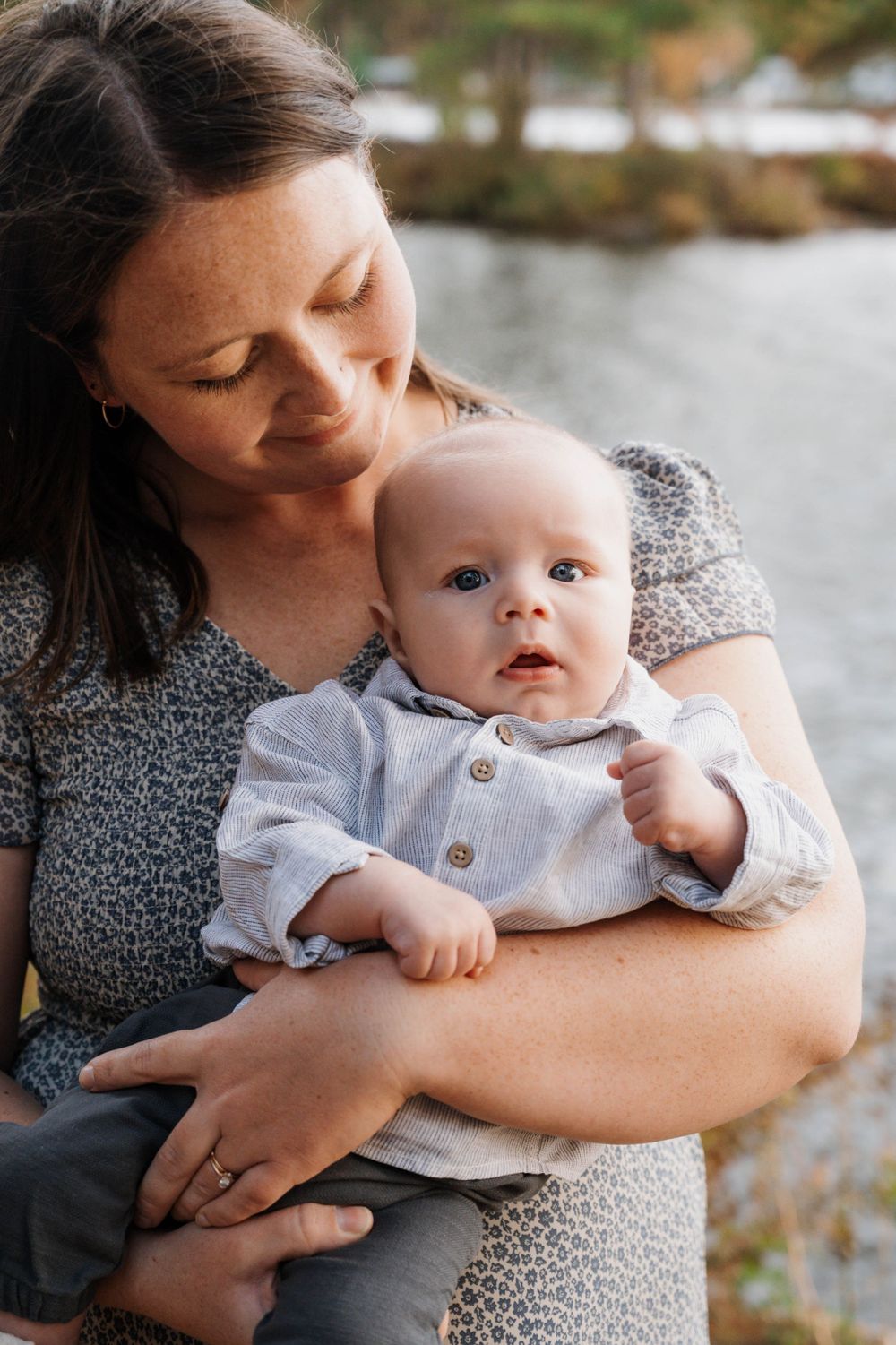 Tender moment as a parent in a grey dress cradles their young baby wearing a light colored outfit near a waterside setting.