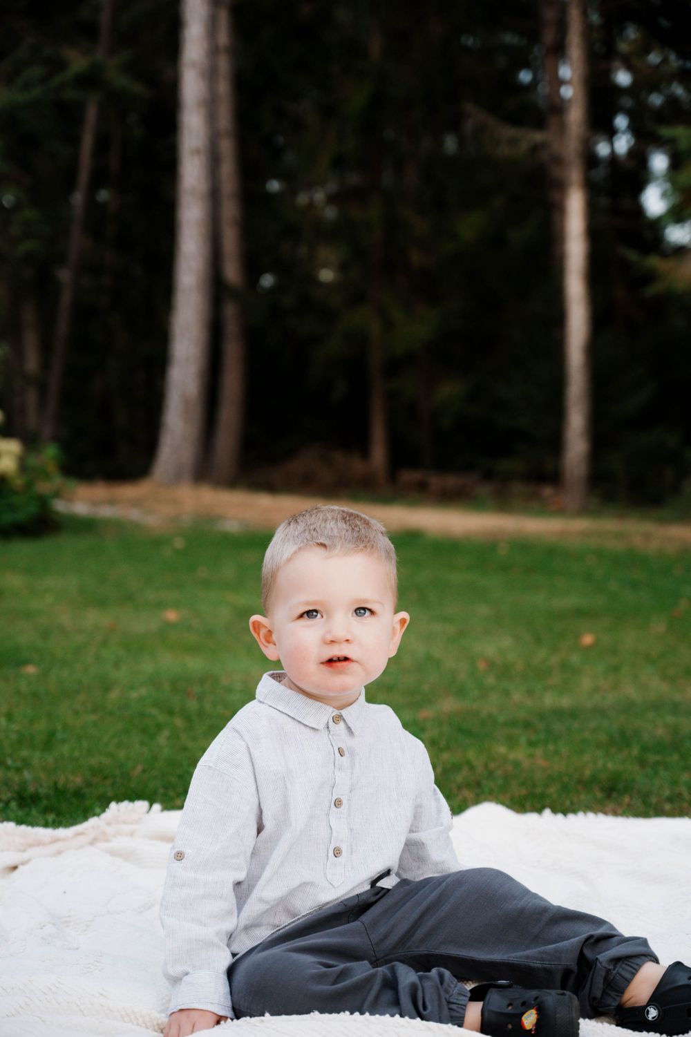 Young child in grey button-up shirt sits on white blanket with forest backdrop.