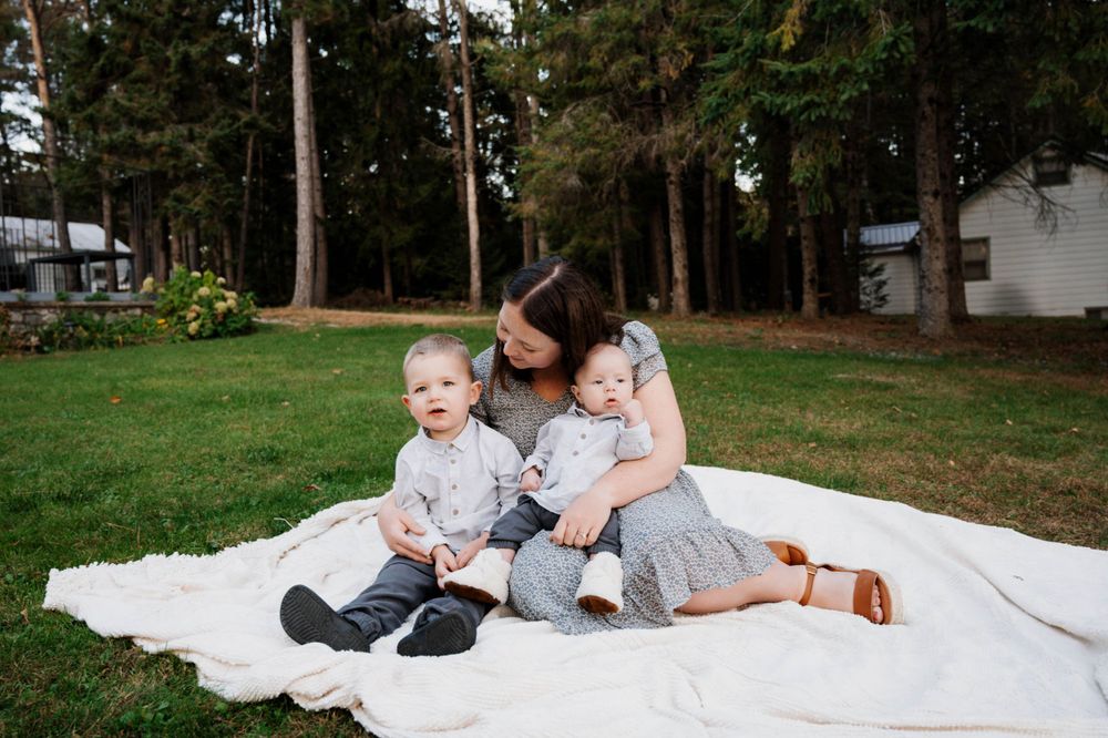 Family sits together on white blanket for outdoor photo session in wooded backyard setting.