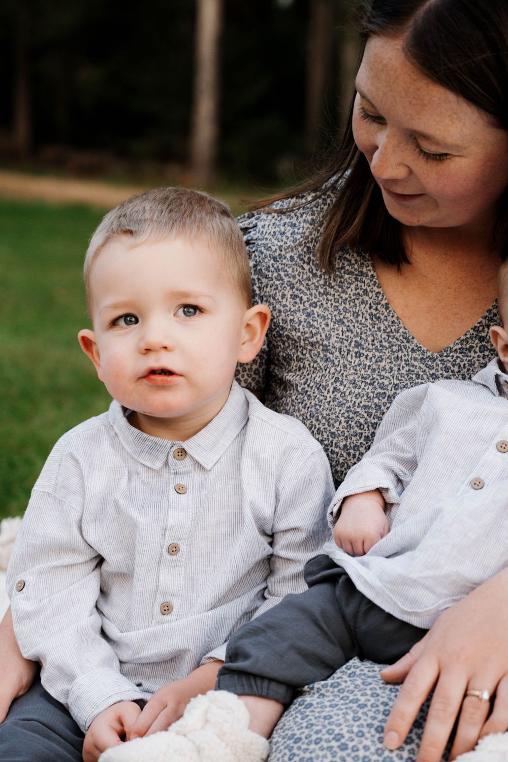 A young child wearing a white button-down shirt sits outdoors with someone in a patterned dress during a family photo session.