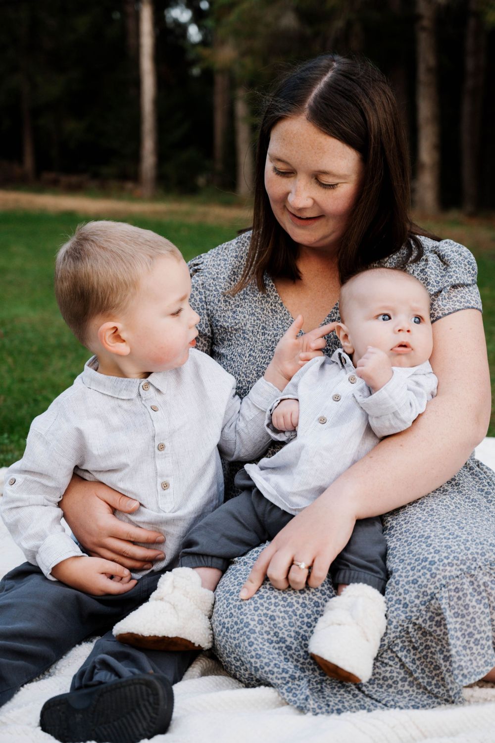 Family sits together outdoors on a blanket, sharing a tender moment in a wooded setting during a casual photo session.