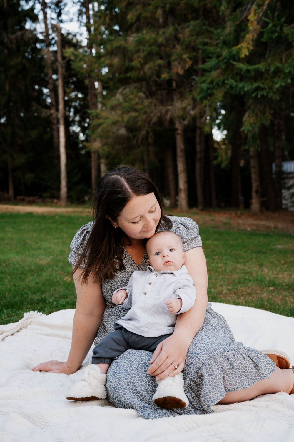 Parent and baby sitting on white blanket outdoors with forest backdrop.