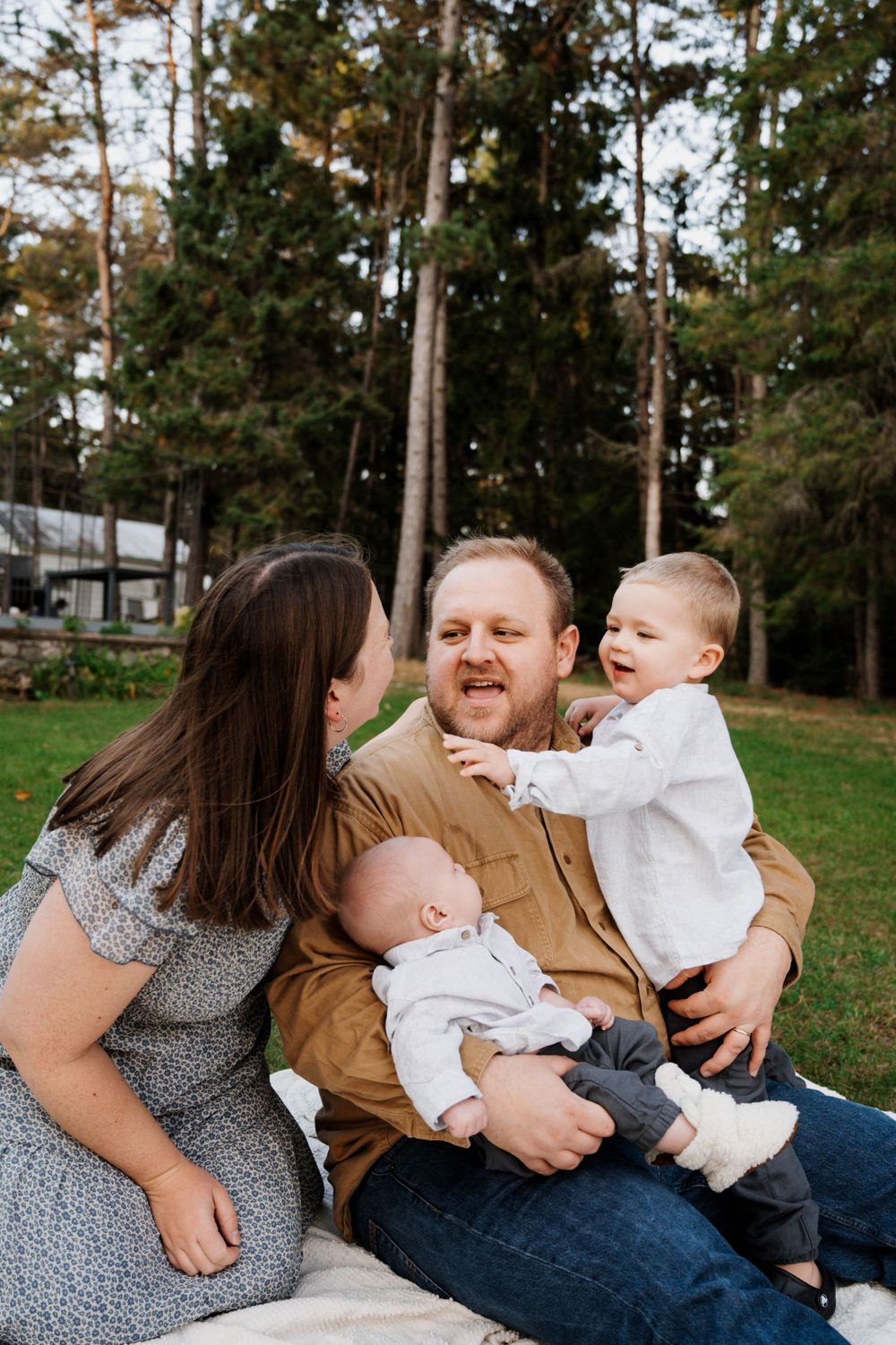 A family sits together on a blanket outdoors with evergreen trees in the background during a photo session.
