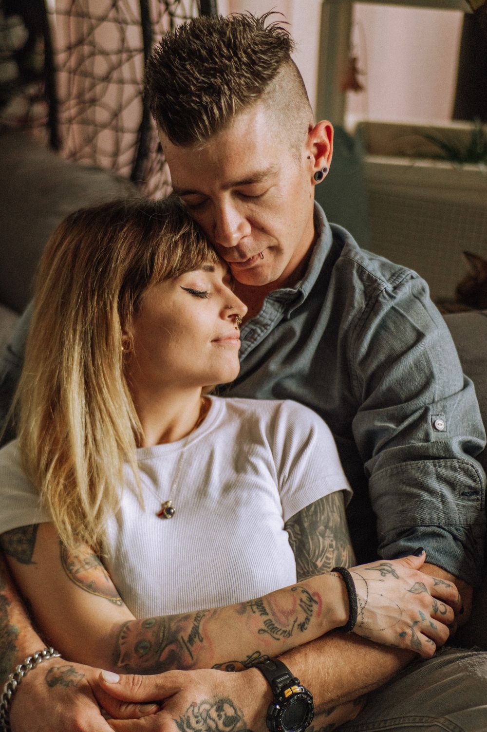 Couple in casual attire sharing tender moment on couch with visible tattoos.