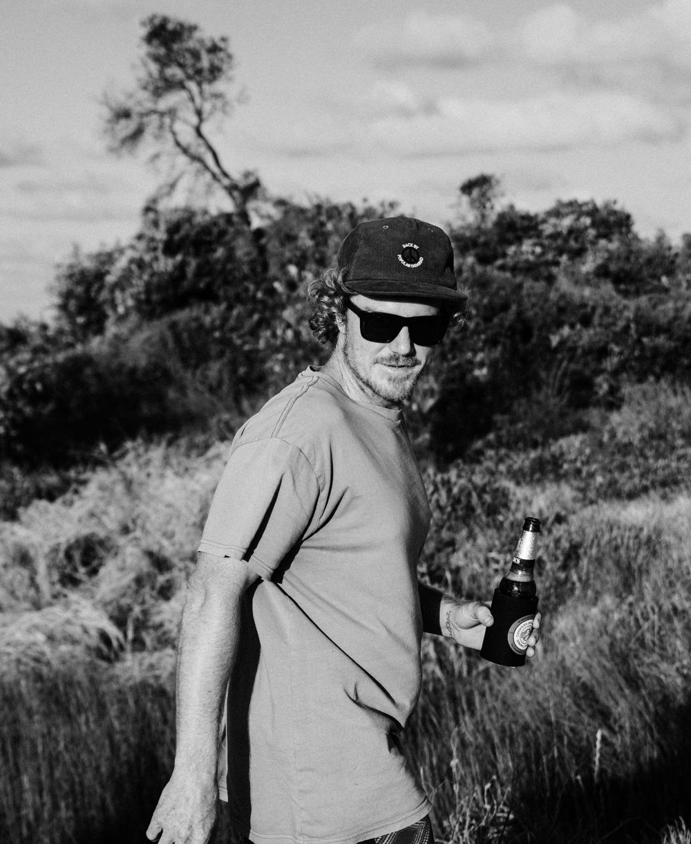 Casual black and white portrait of someone in sunglasses and cap holding a drink outdoors.