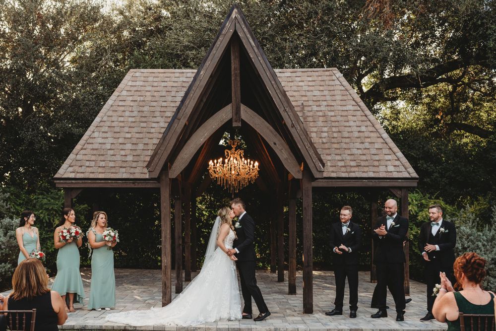 Wedding ceremony taking place under rustic wooden chapel with wedding party standing at altar.