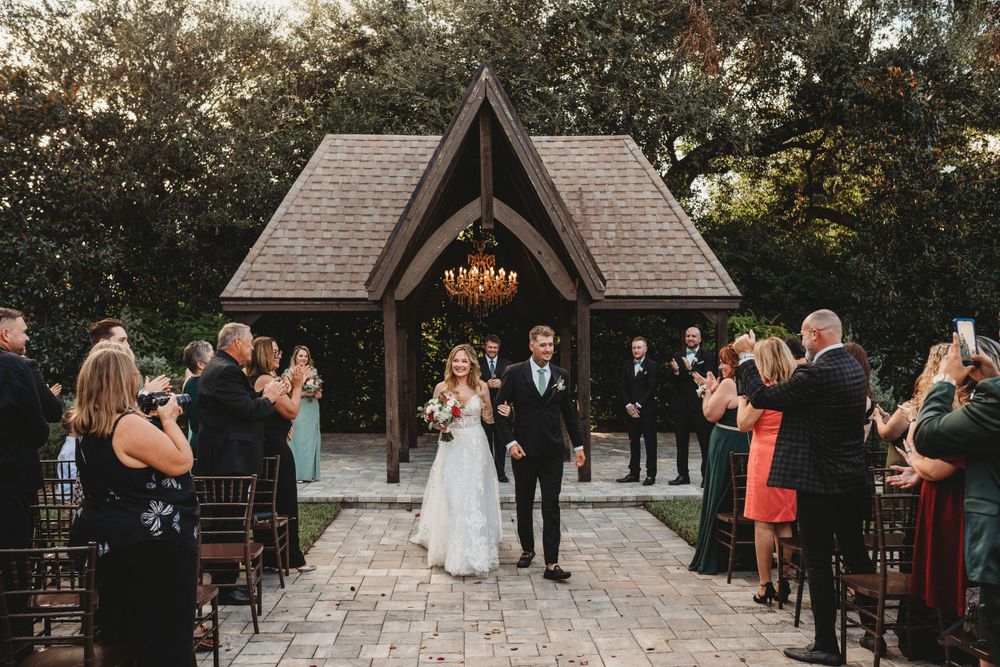 Wedding party exits rustic wooden chapel with vaulted ceiling and chandelier while guests cheer on stone pathway.