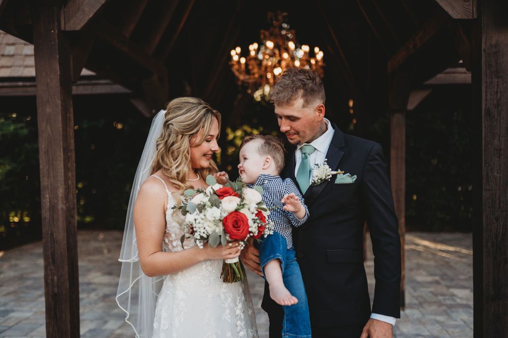 White floral bouquet complements the elegant bride and groom holding baby at outdoor wedding venue.