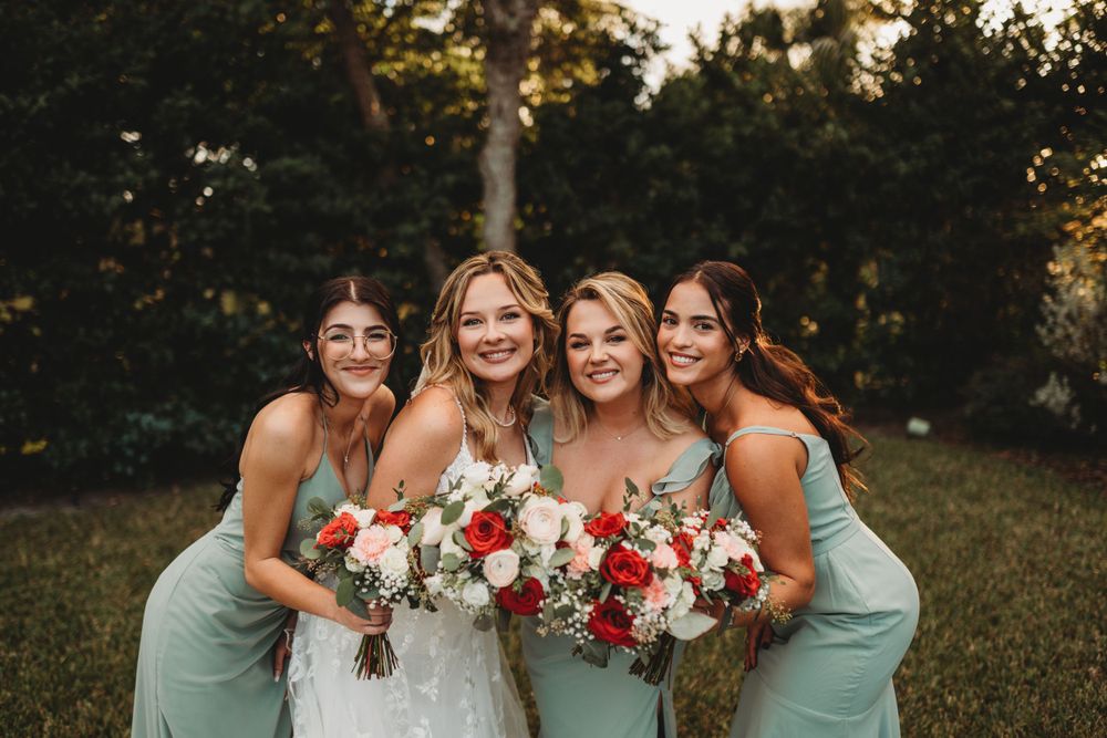 Wedding party laughs together while holding vibrant red and white bouquets against sunset backdrop.