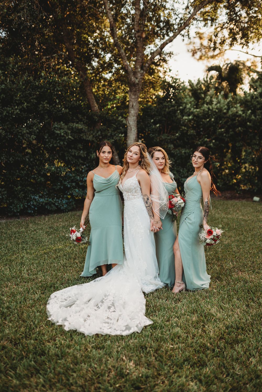 Bridal party in mint green dresses poses on grass with lace wedding gown train in evening light.