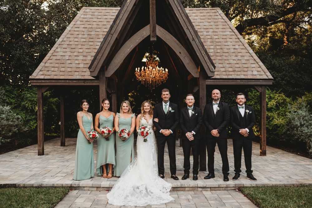 Wedding party poses formally in front of wooden chapel with elegant chandelier and stone pathway.