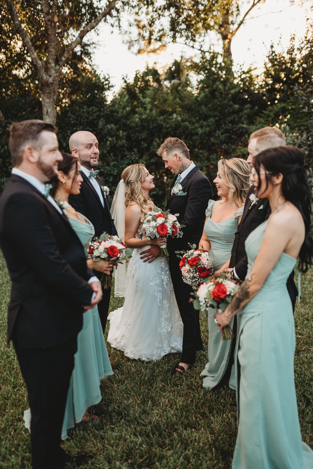 Wedding party laughs together outdoors with bridesmaids in mint dresses holding red and white bouquets.
