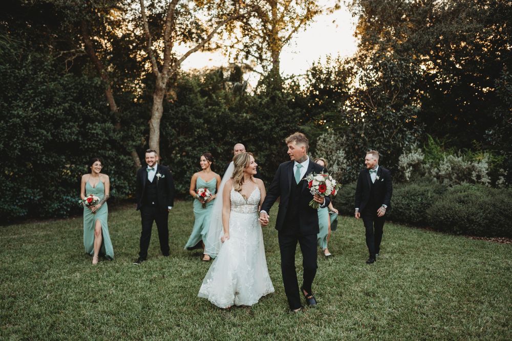 Wedding party walks across green lawn with rustic chapel and trees in background at sunset.