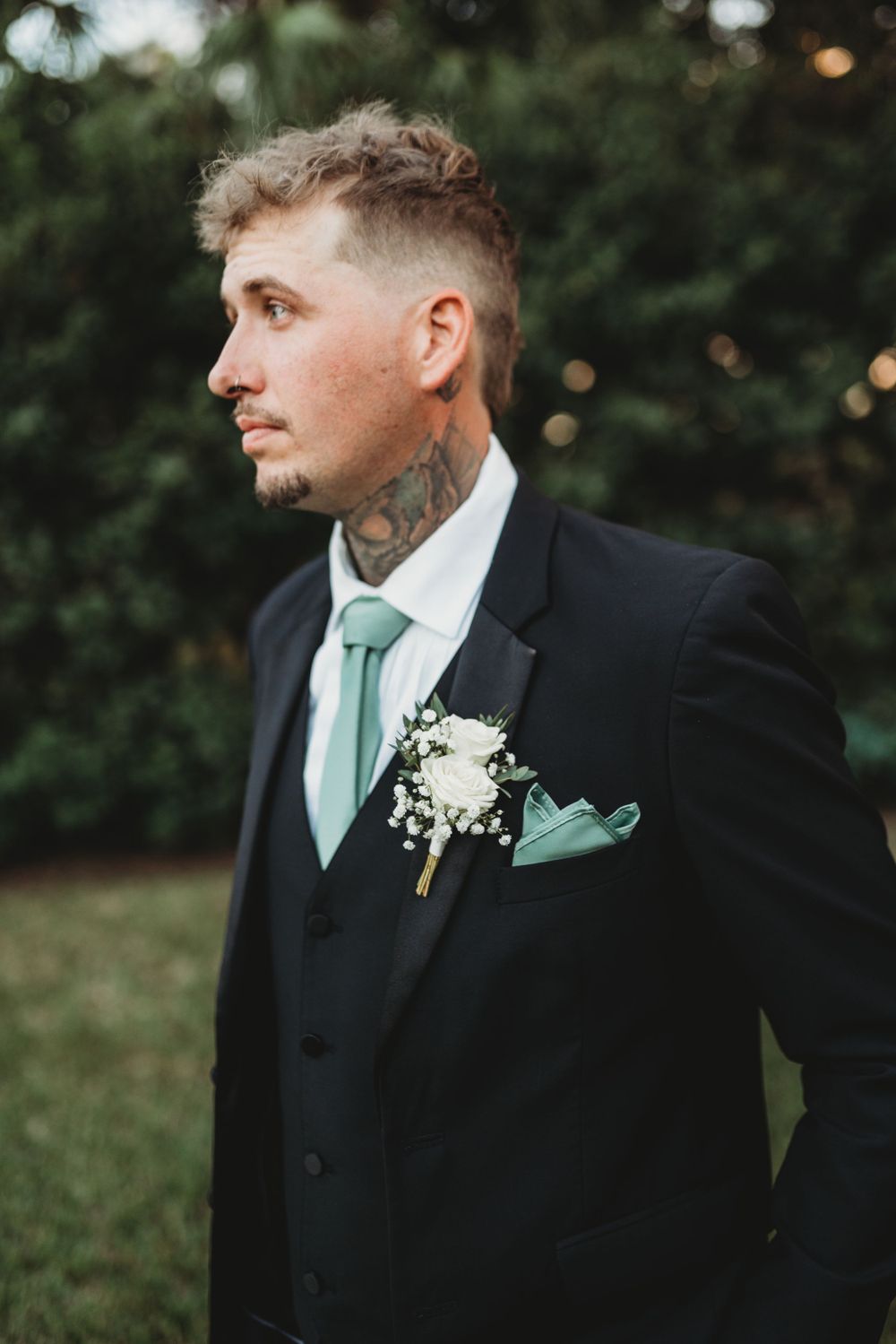 Groom in black suit with mint green tie and floral boutonniere looking to the side outdoors.