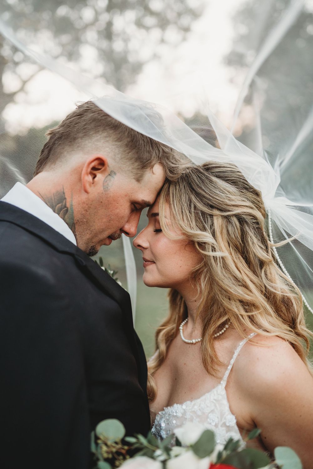 Intimate moment between newlyweds with bride's veil flowing in outdoor setting.