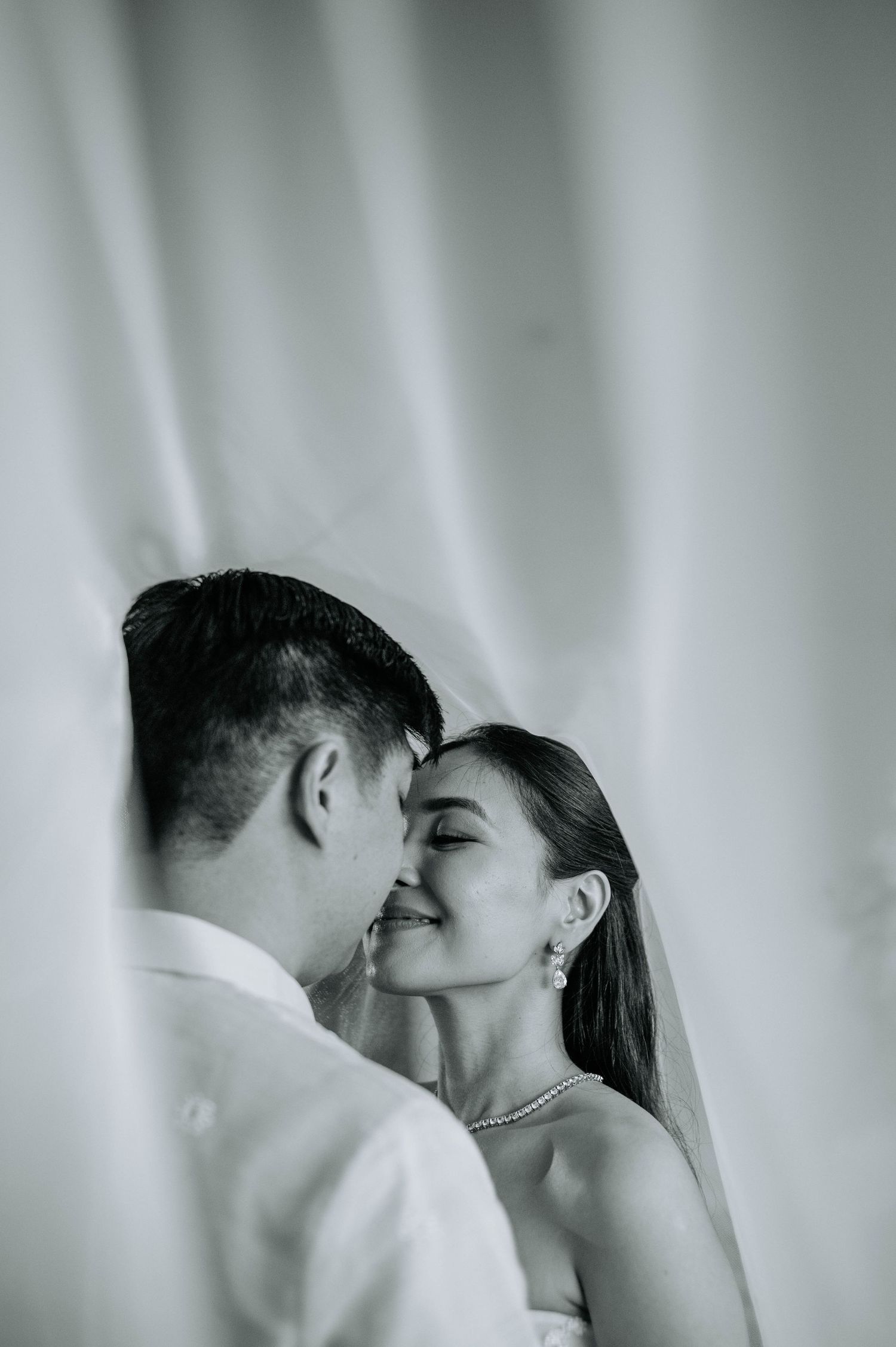 Intimate black and white wedding portrait with soft lighting and curtain backdrop.