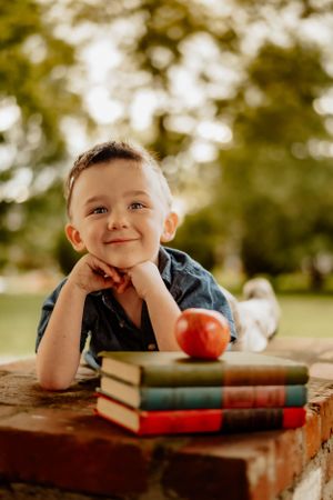 Young student rests head on hands while lying next to stack of books outdoors.