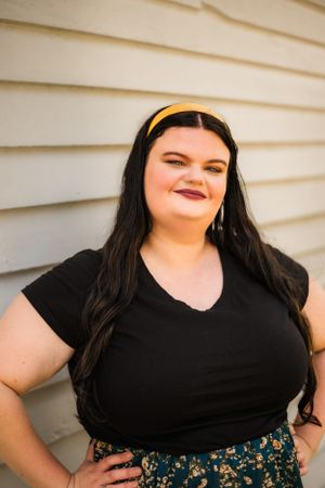 Young woman in black shirt and yellow headband poses confidently against white wall.