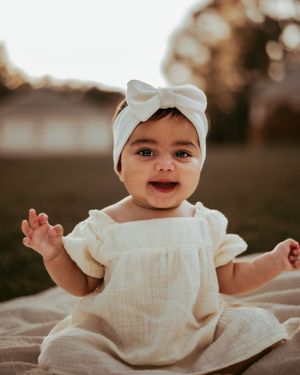 Smiling baby in white dress and bow headband sits outdoors at sunset.