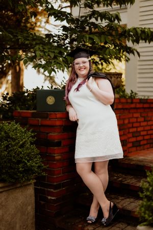 Graduate in white dress poses with diploma holder by brick wall and greenery.