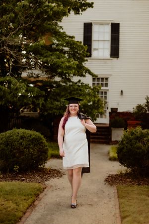 Graduate in white dress and mortarboard walking along pathway in front of white building.