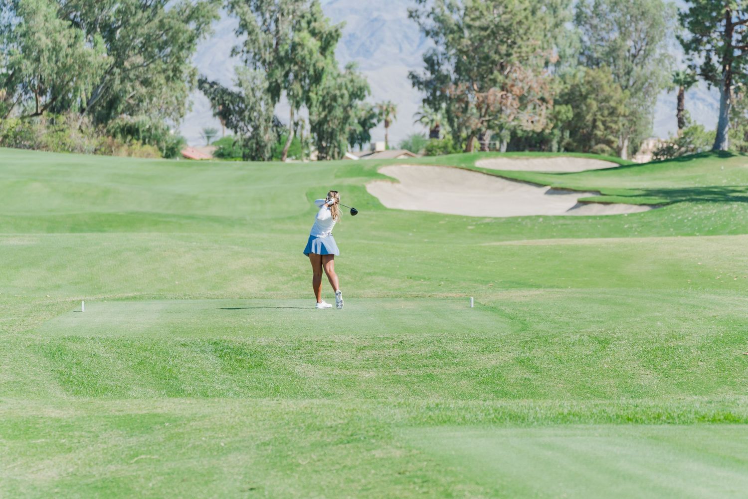 A golfer stands on a lush green course with sand bunkers and trees visible in the background on a sunny day.