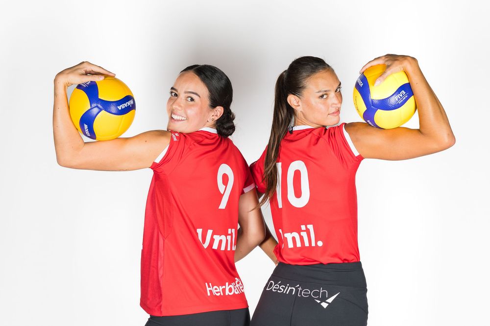 Two volleyball players in red jerseys numbered 9 and 10 pose back to back while holding volleyballs.
