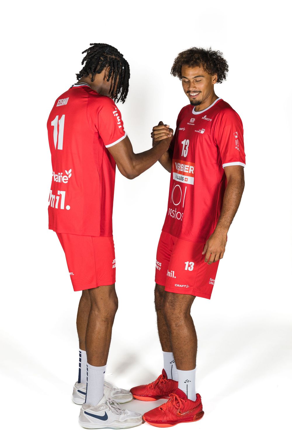 Two athletes in red sports uniforms and sneakers shake hands while standing against a white background.