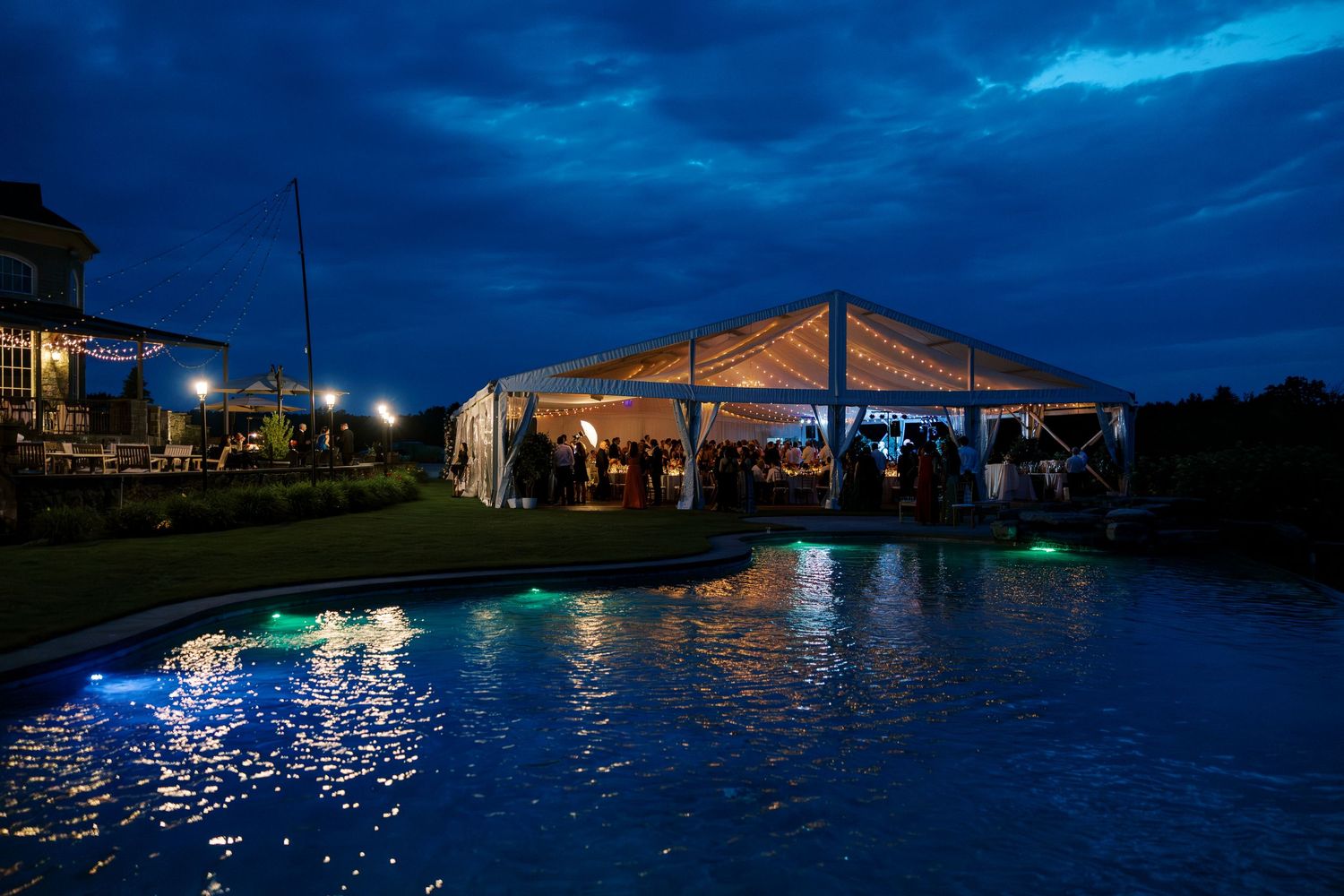 An illuminated event tent beside a pool reflects beautifully in the calm water during twilight hour.