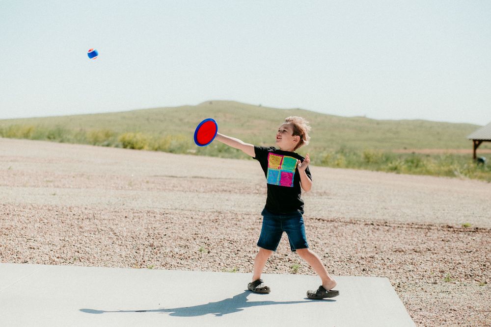 Child in a sun hat plays with a red frisbee on a dirt road with desert hills in the background.
