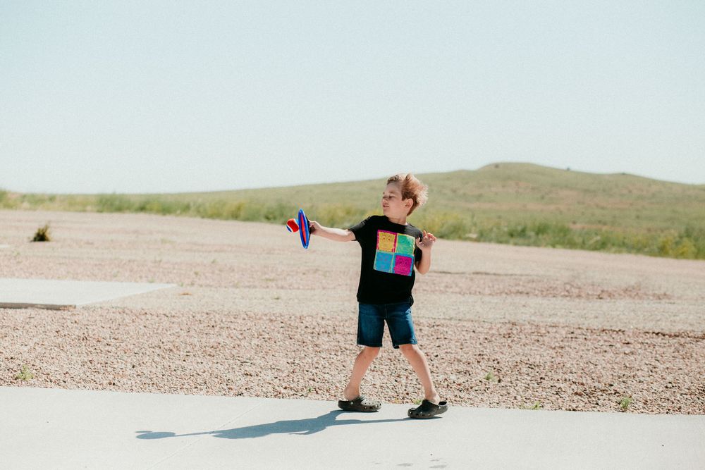 A person wearing black shorts and a backpack stands with outstretched arms on a desert path surrounded by hills.