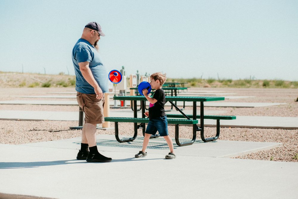 Friends enjoying multiple games of table tennis on green outdoor ping pong tables in an arid setting.