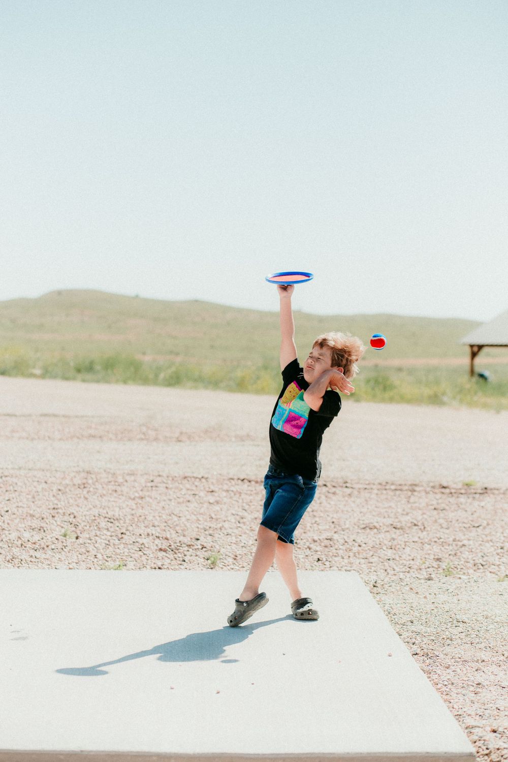 A person in a black t-shirt and shorts throws a frisbee while standing on a concrete pad in a desert landscape.