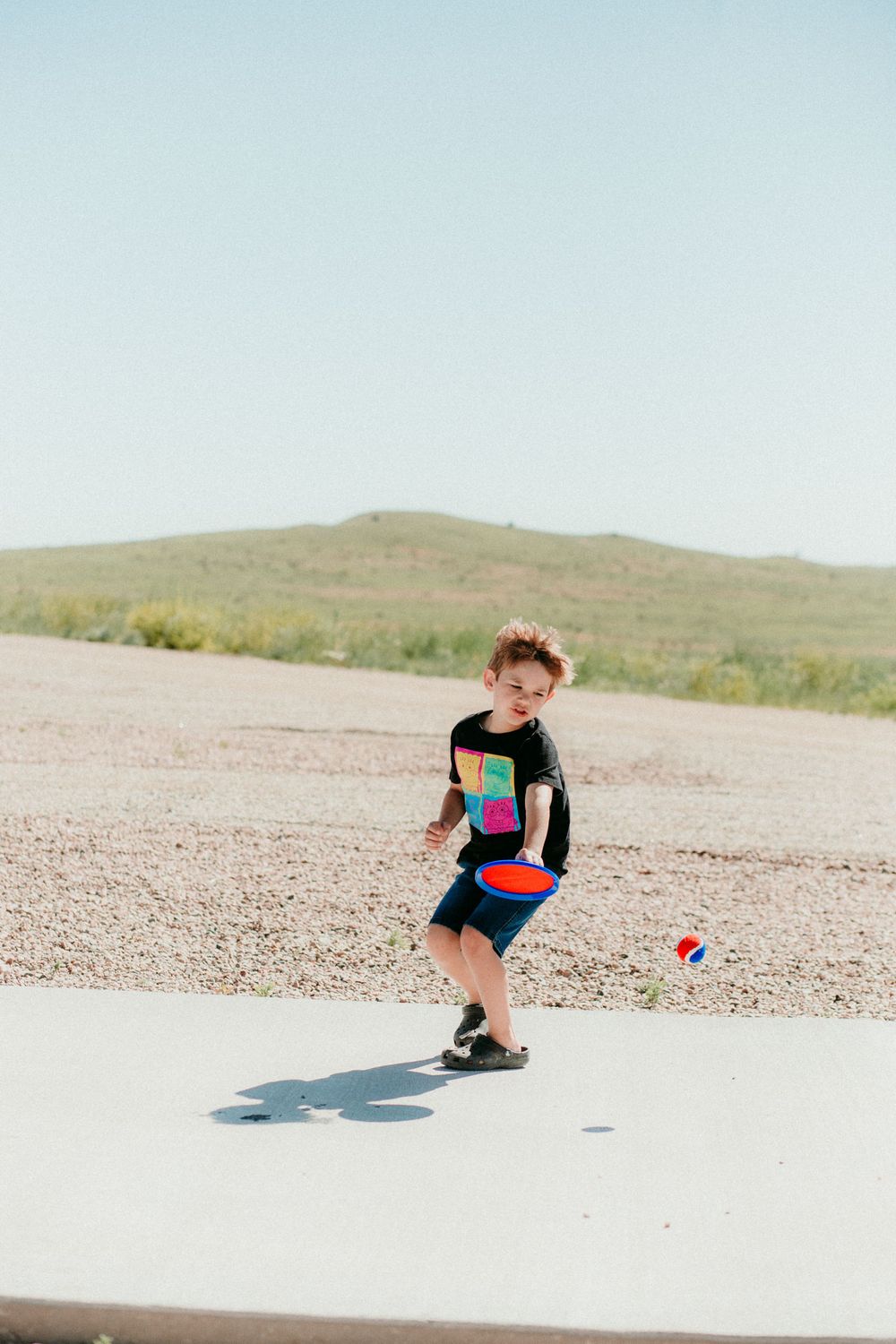A child in a colorful swimsuit plays with a ball on a bright, sandy beach on a sunny day with rolling hills in the background.