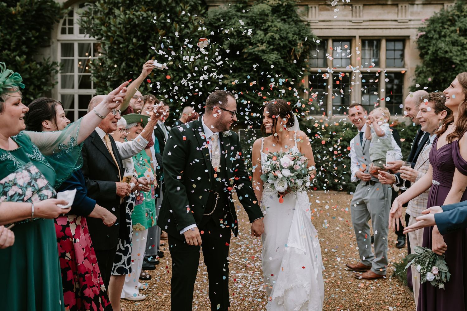 Wedding guests throw white confetti over newlyweds during outdoor ceremony recessional at elegant country estate.